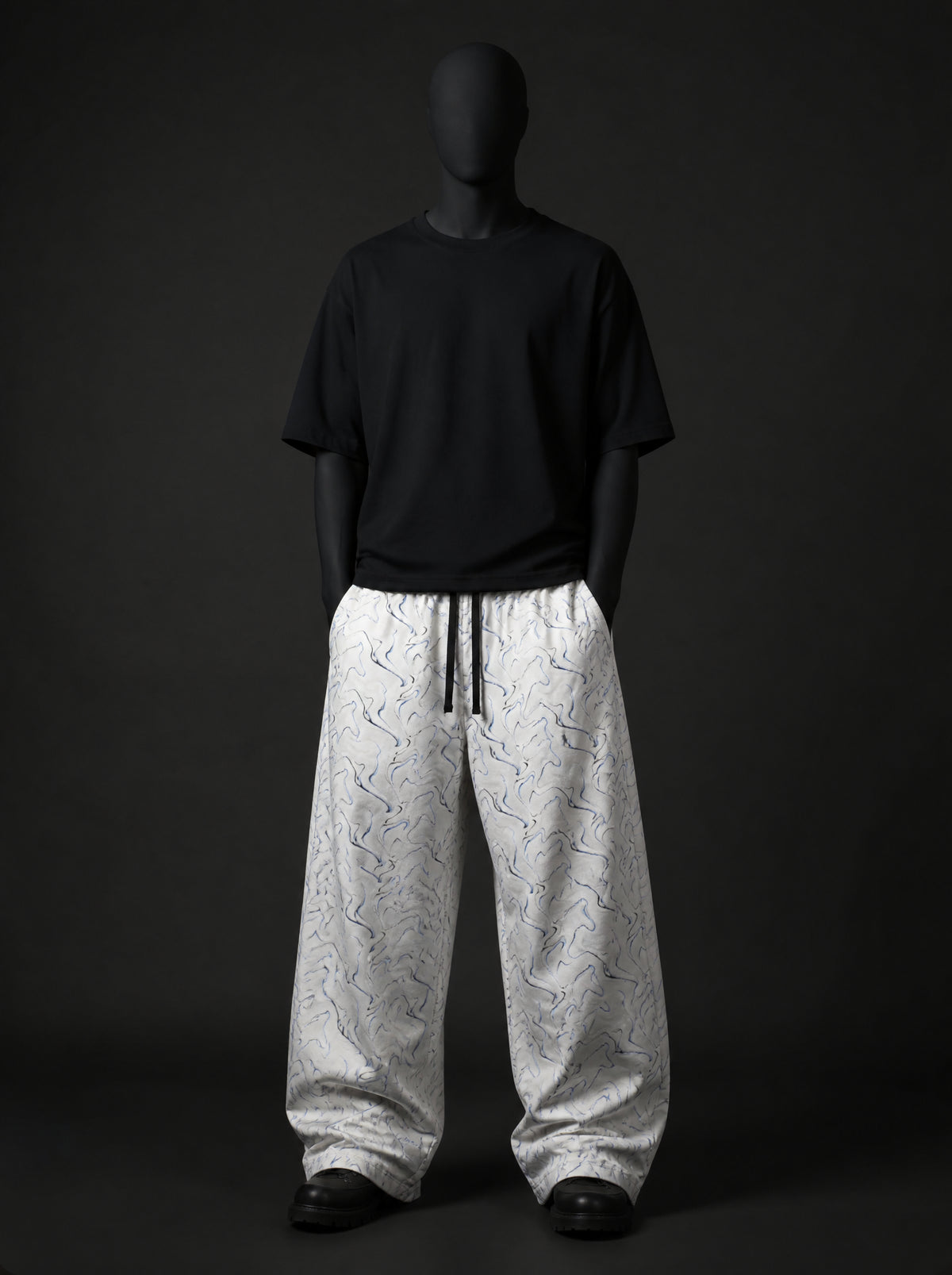 Full-length front view of white swirl-print baggy pants with black drawstring, styled with black t-shirt and boots