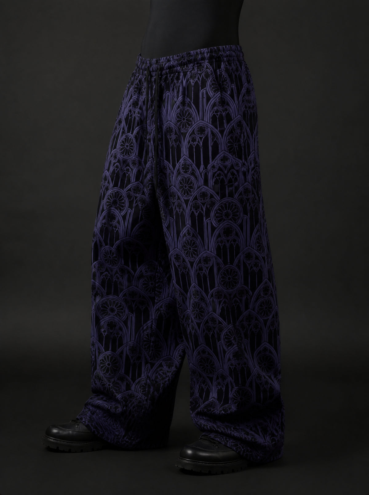 Three-quarter side view of baggy purple gothic arch print pants with drawstring waist, worn with black boots