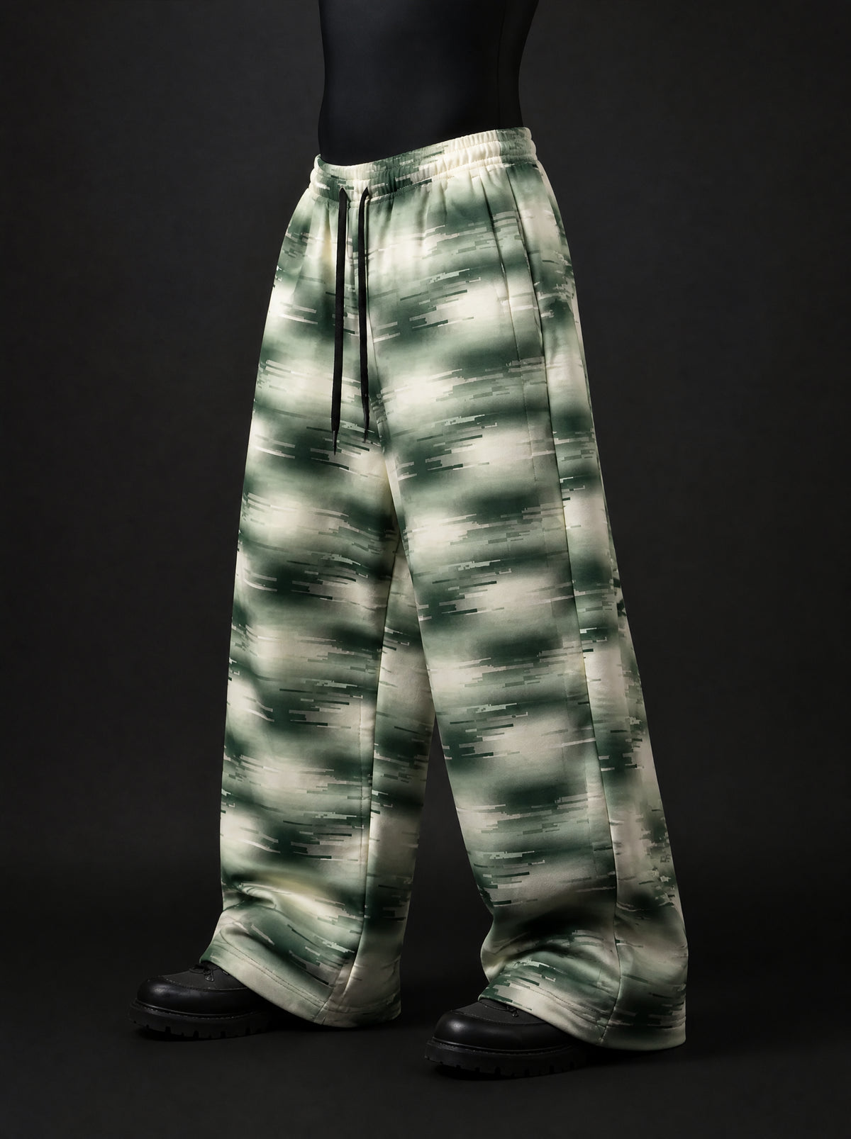 Three-quarter view of baggy green and cream glitch-print trousers with black drawstring waist and black lug-sole boots