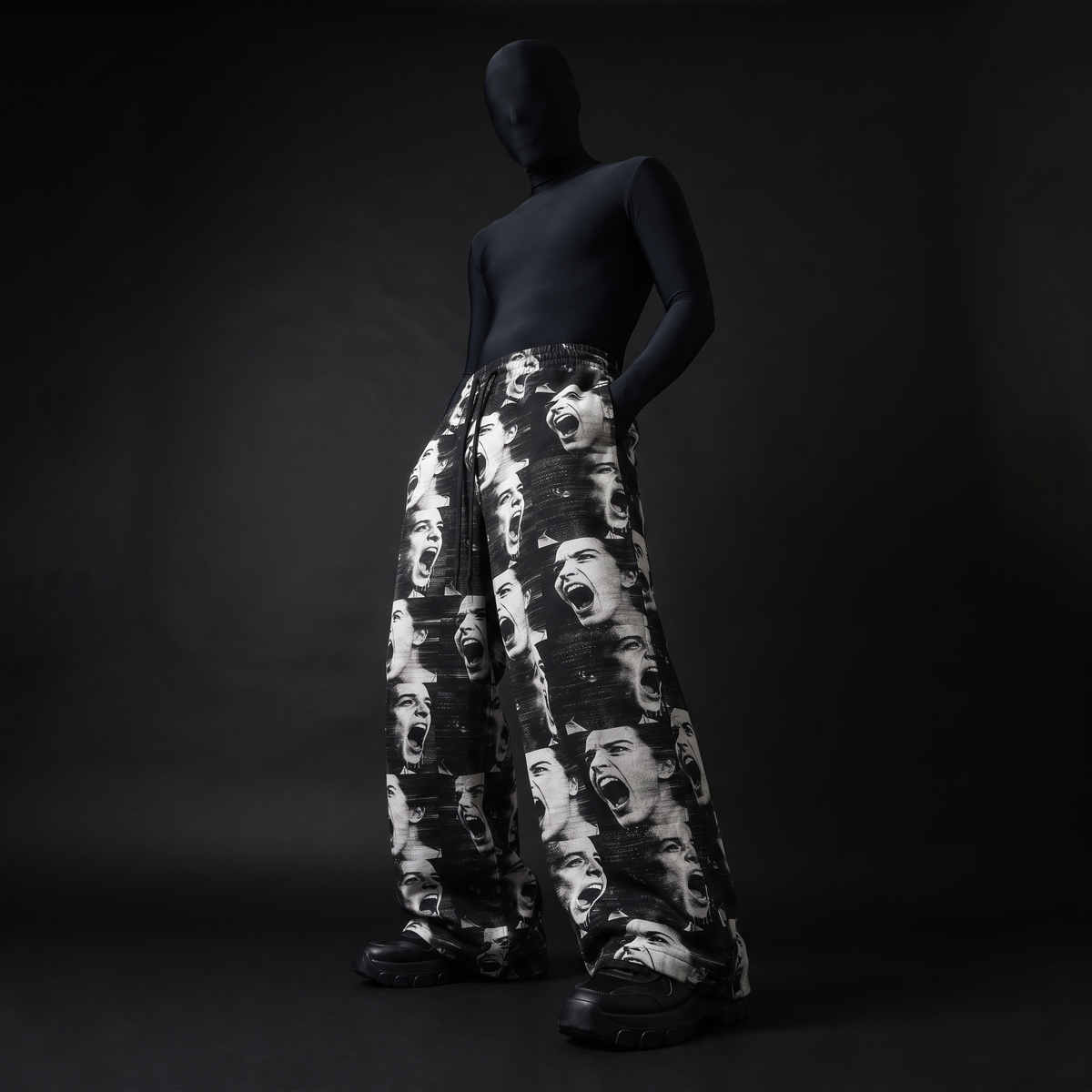 Model in black bodysuit and mask wearing wide-leg pants with black-and-white screaming faces print, hand in pocket